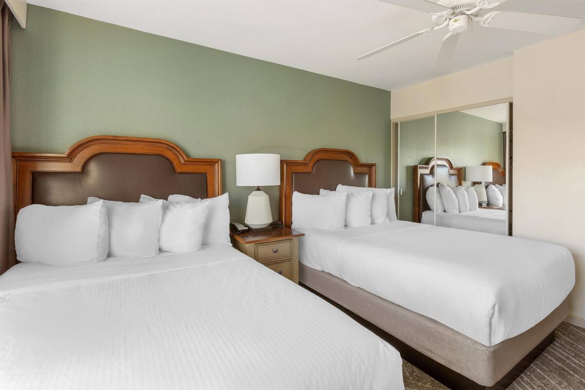 A hotel room with two beds, a nightstand with a lamp, a ceiling fan, and a large mirror on the wall.