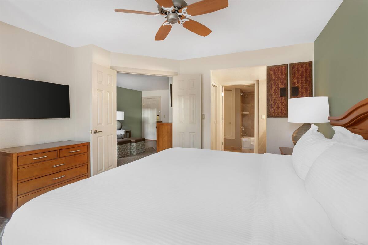A hotel room with a large bed, a wooden dresser, a ceiling fan, and a flat-screen TV mounted on the wall.