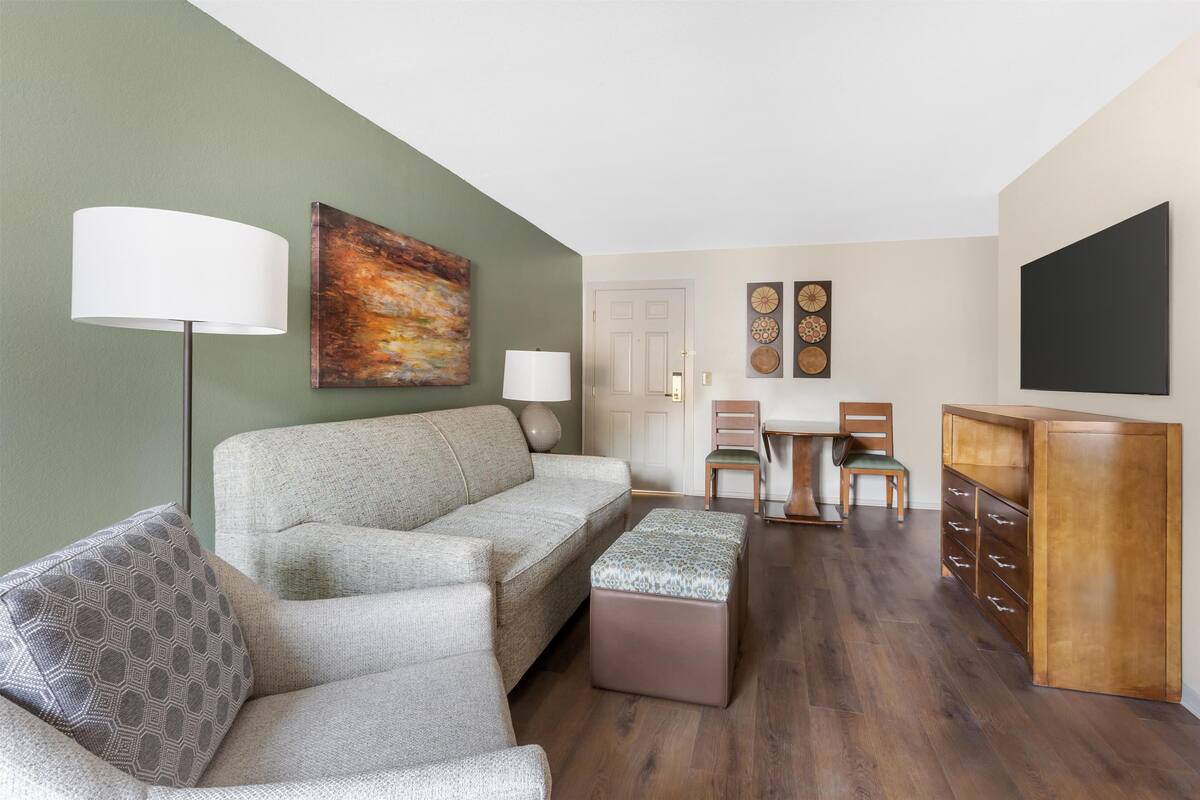 A living room with a sofa, ottoman, and a coffee table. There is a lamp, a painting, and a TV on the wall.