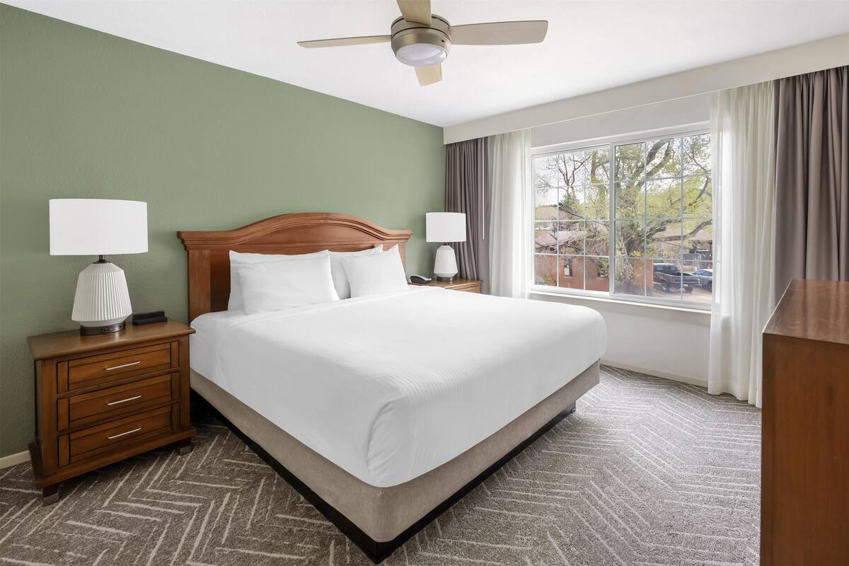A hotel room with a large bed, two bedside tables with lamps, a ceiling fan, and a view of the outside through a window.