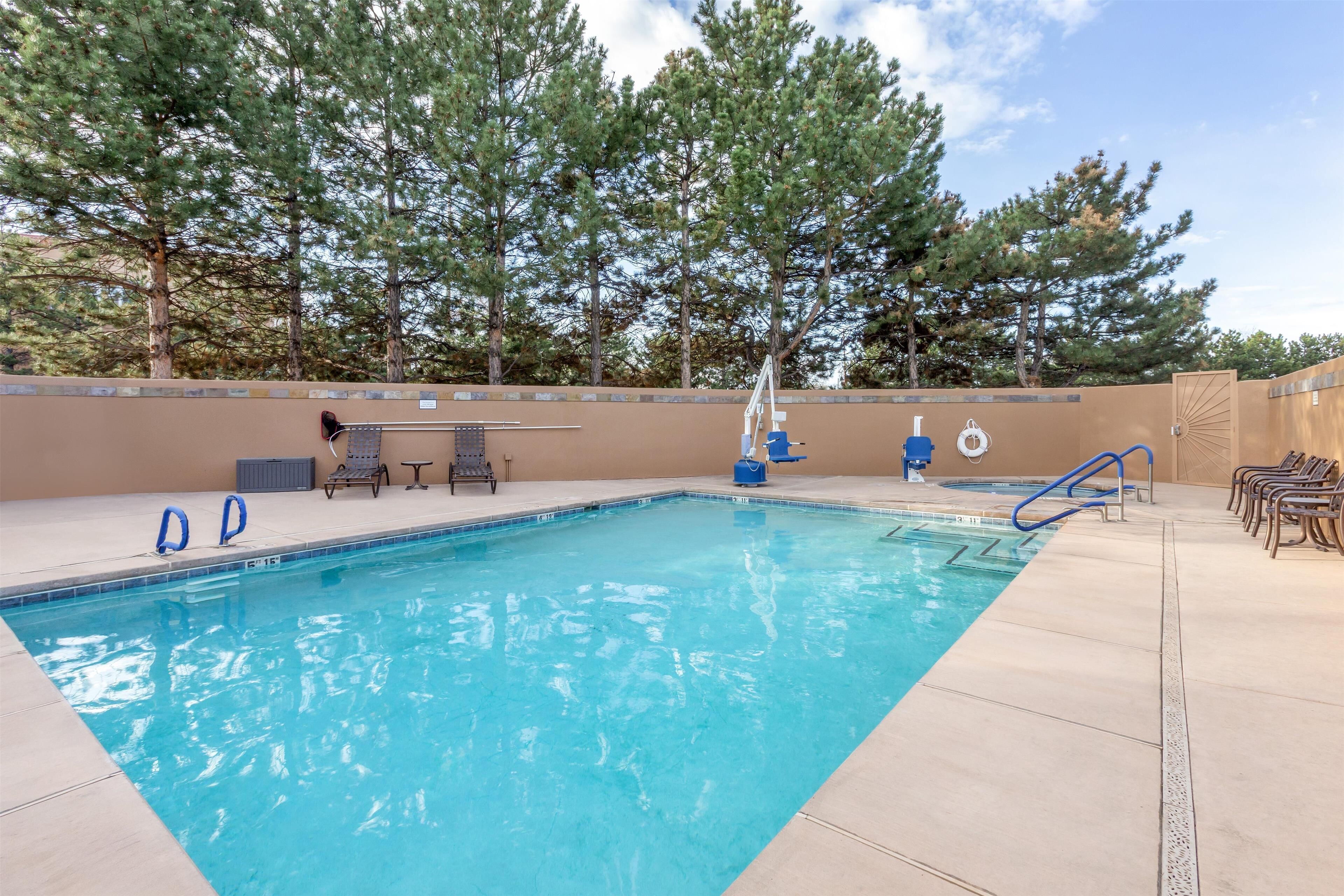 A rectangular swimming pool with clear blue water, surrounded by a beige tiled deck. There are lounge chairs and a small table on the deck.