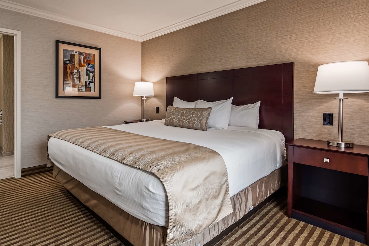 A hotel room with a large bed, a nightstand with a lamp, a framed artwork on the wall, and a bathroom with a toilet.