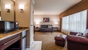 A hotel room with a kitchenette, a living area with a sofa and ottoman, a dining table, and a large window with curtains.
