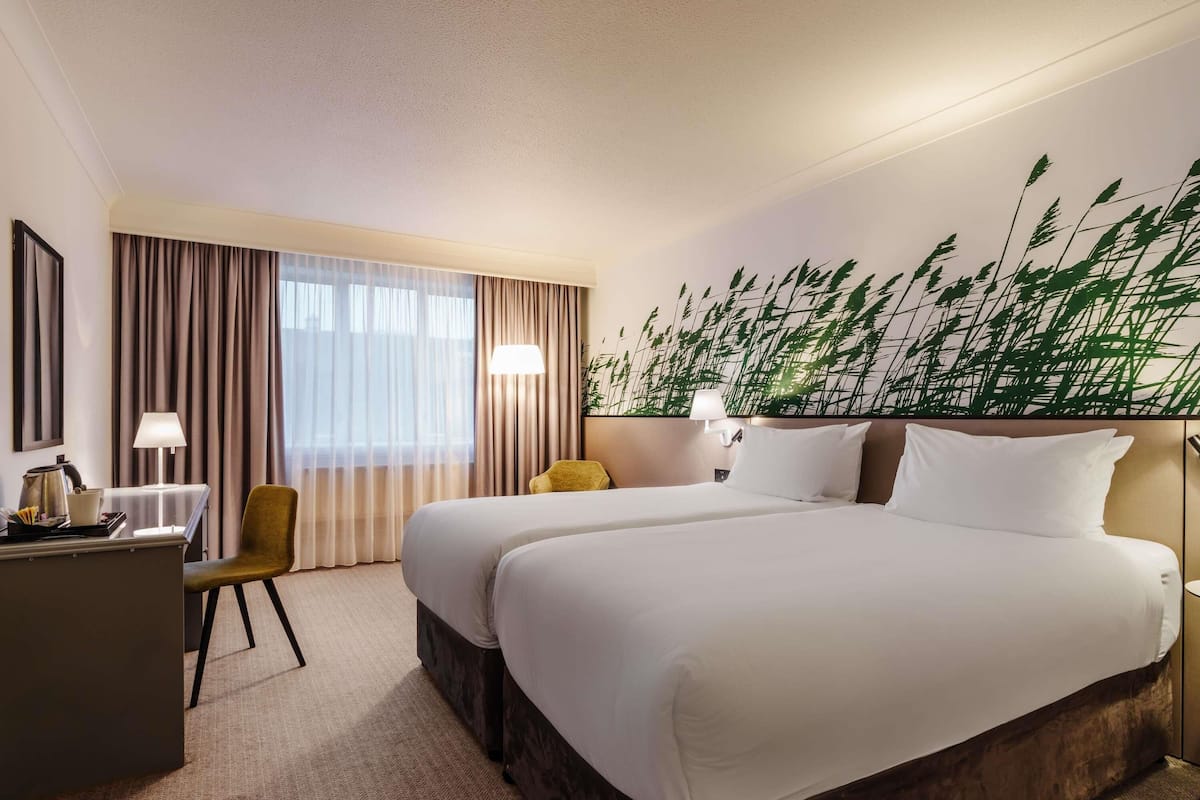 Habitación de hotel con dos camas, un escritorio, una silla, una lámpara y un mural grande en la pared.
