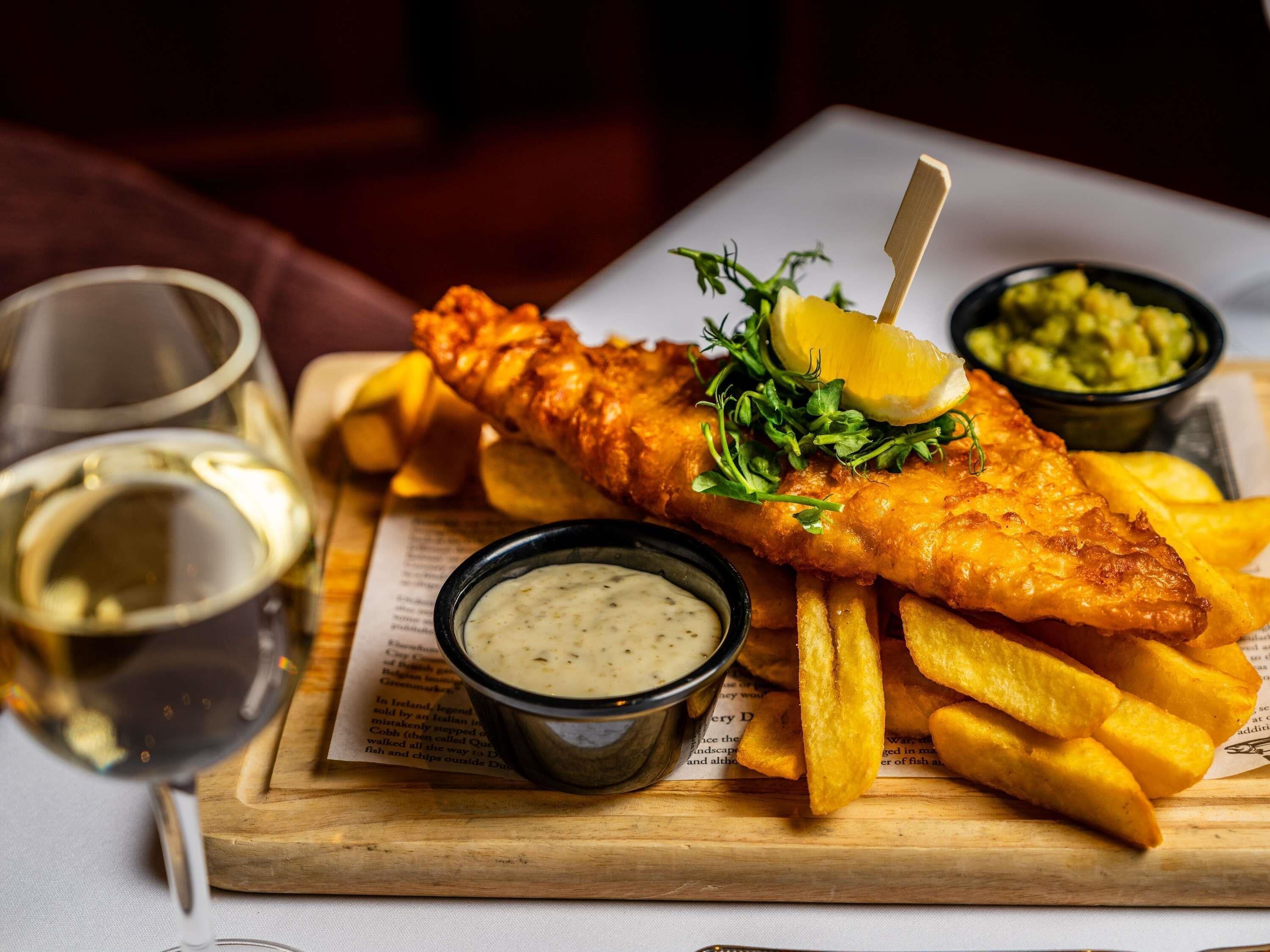 A meal of fish and chips with two dipping sauces and a lemon wedge on a wooden board.
