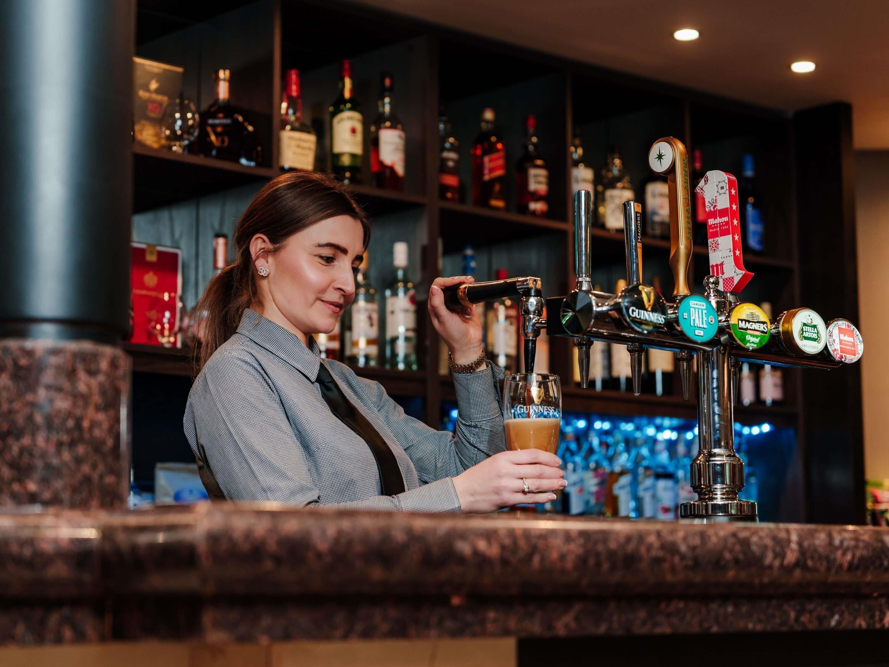 Un bartender sirve una bebida de un grifo con la etiqueta 'GUINNESS' en un bar con una variedad de botellas de licor al fondo.