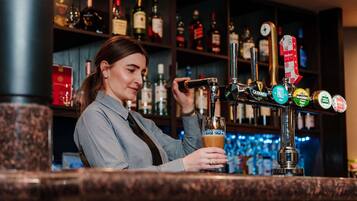 Un bartender sirve una bebida de un grifo con la etiqueta 'GUINNESS' en un bar con una variedad de botellas de licor al fondo.