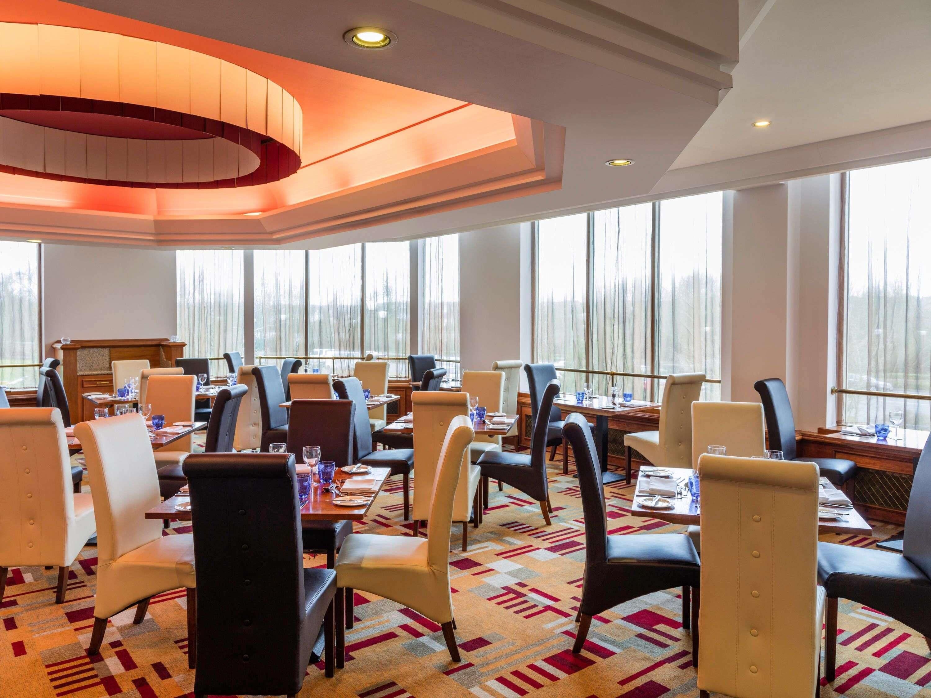 A hotel dining area with multiple tables and chairs, large windows, and a patterned carpet.