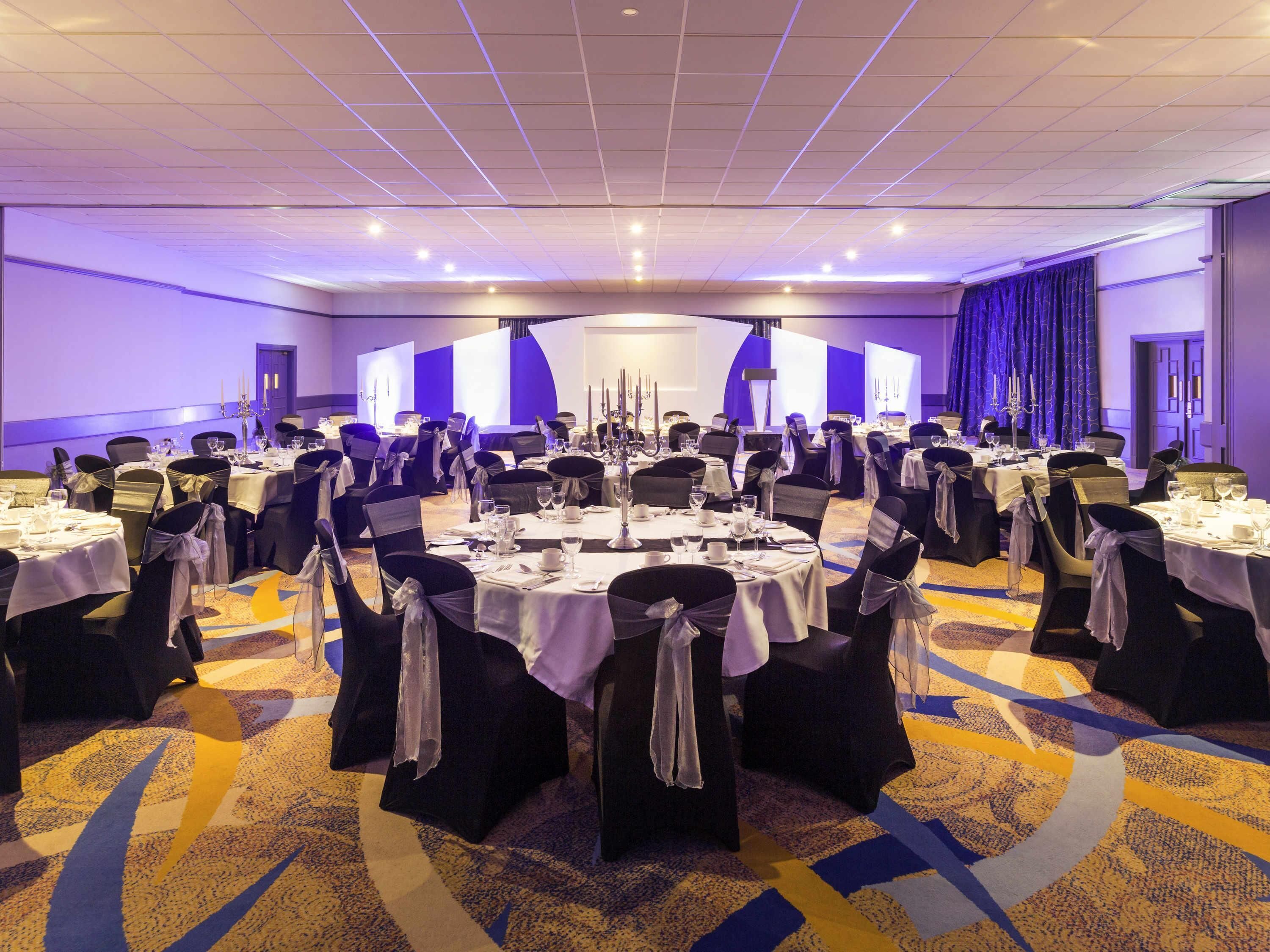 A large banquet hall with round tables, black chairs, and white tablecloths.