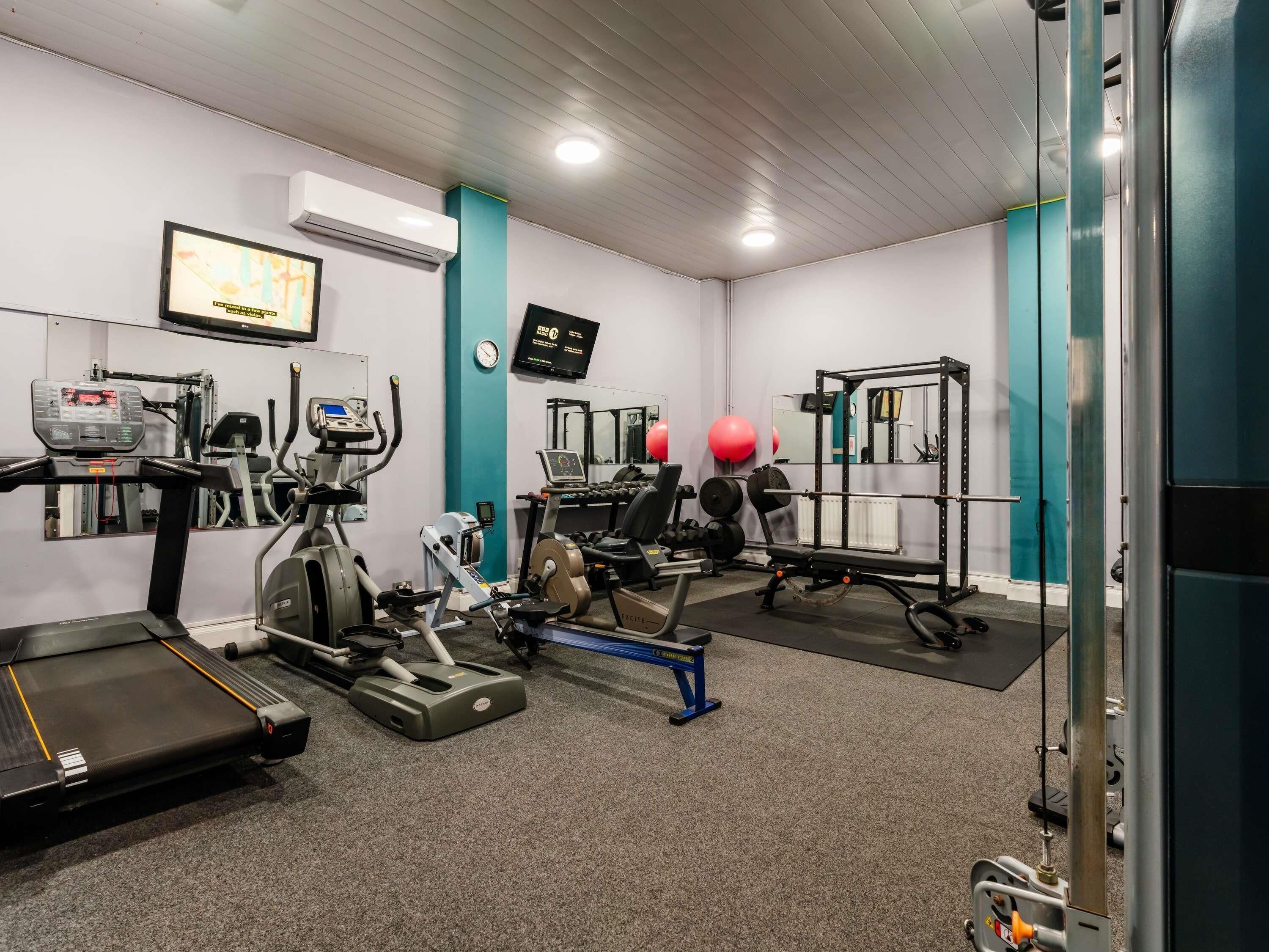 Ein Fitnessraum mit Trainingsgeräten, Laufband, Kraftmaschinen und einem an der Wand montierten Fernseher.