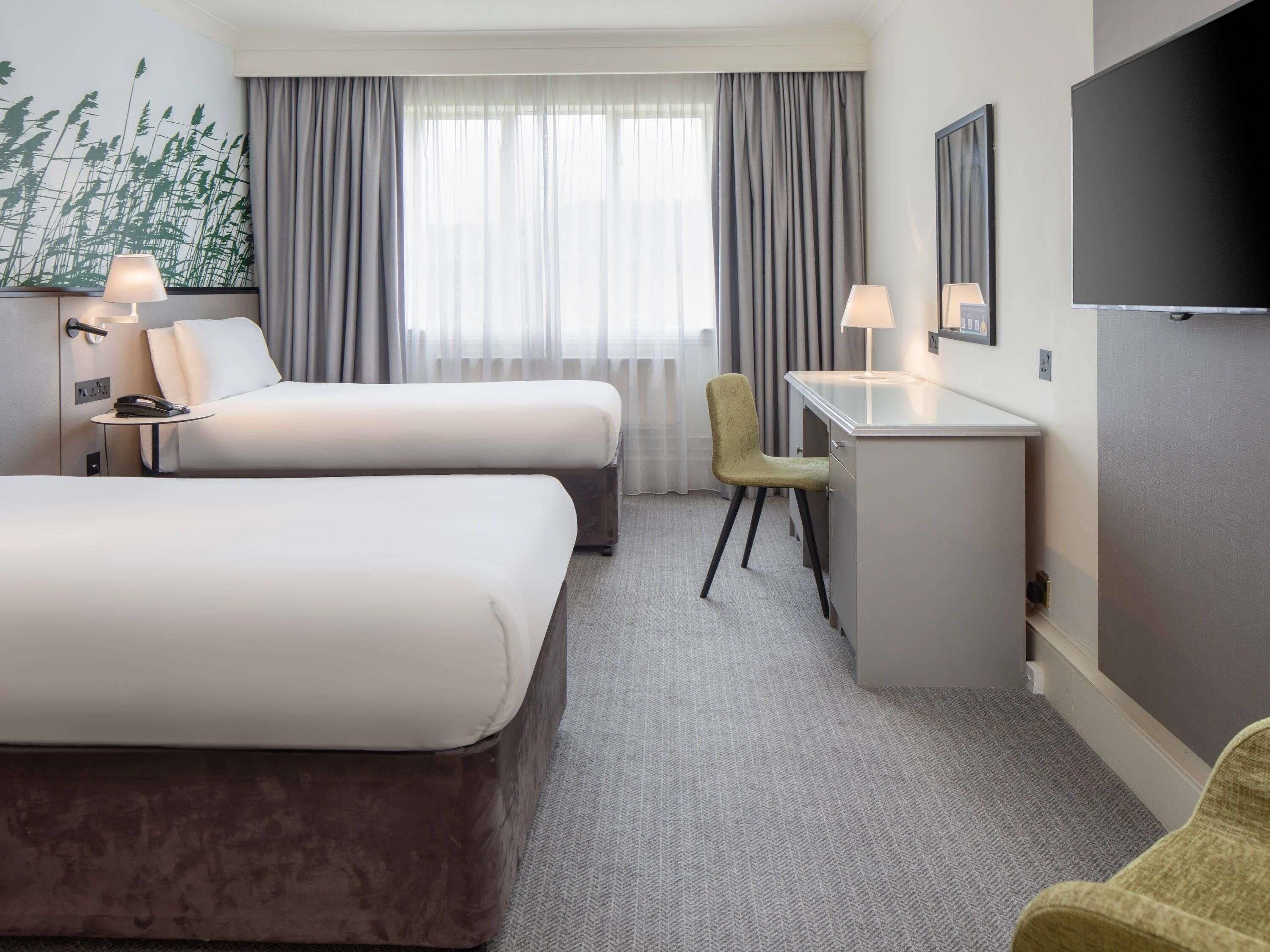 A hotel room with two beds, a desk, a chair, a TV, and a window with curtains.