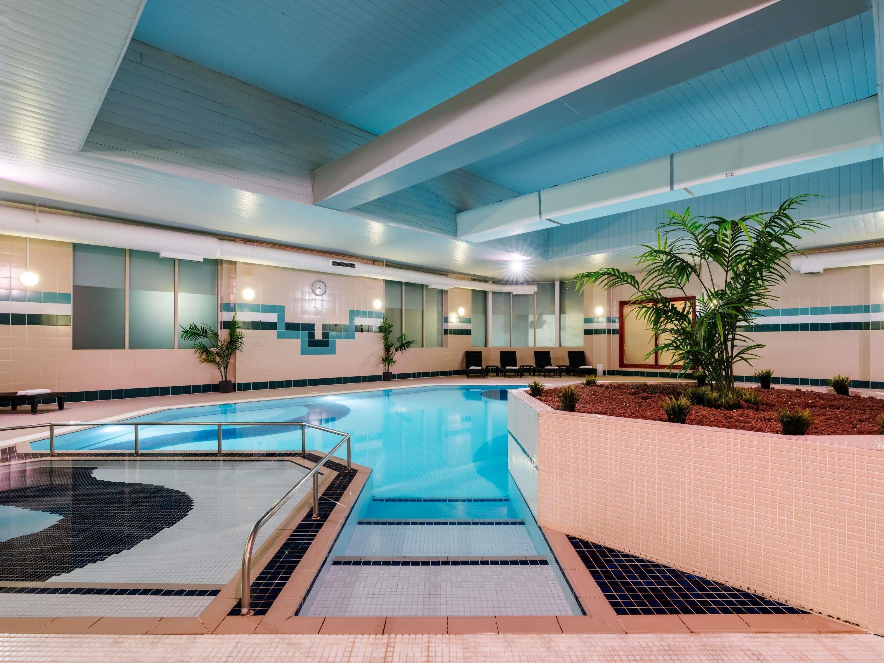 An indoor swimming pool with tiled walls, a clock, and lounge chairs.
