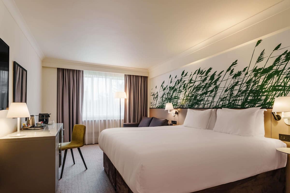 Una habitación de hotel con una cama grande, un escritorio con lámpara, una silla, una mesita de noche con lámpara y un mural de plantas en la pared.