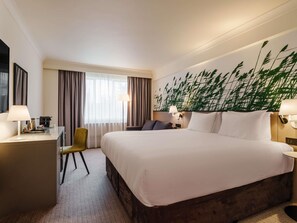 Quarto de hotel com uma cama grande, uma mesa com luminária, uma cadeira, mesa de cabeceira com luminária e um mural de planta na parede.