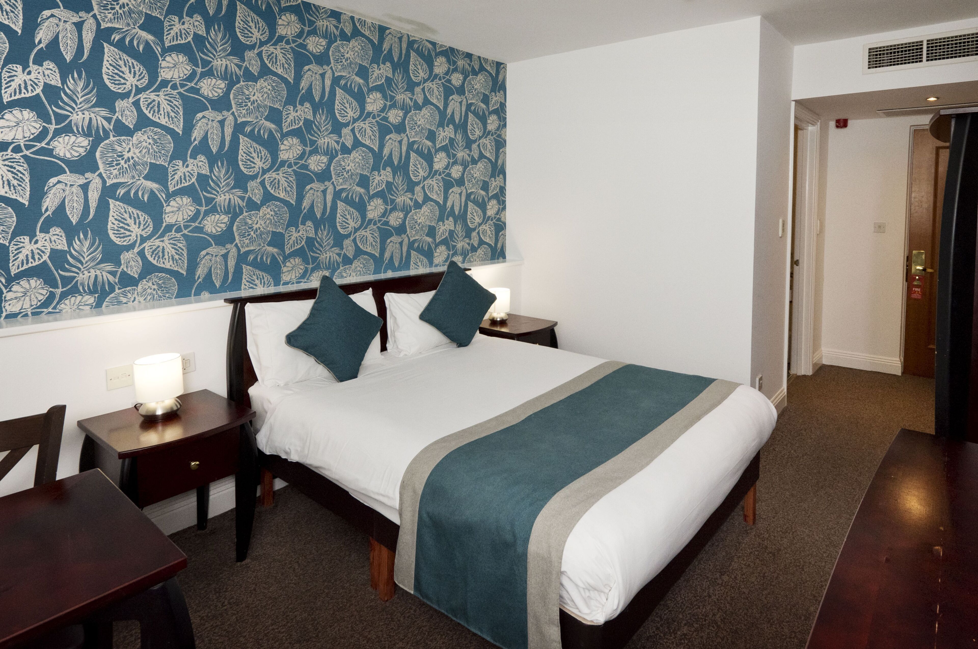 A hotel room with a bed, a nightstand, a table, and a wall with a leaf pattern.