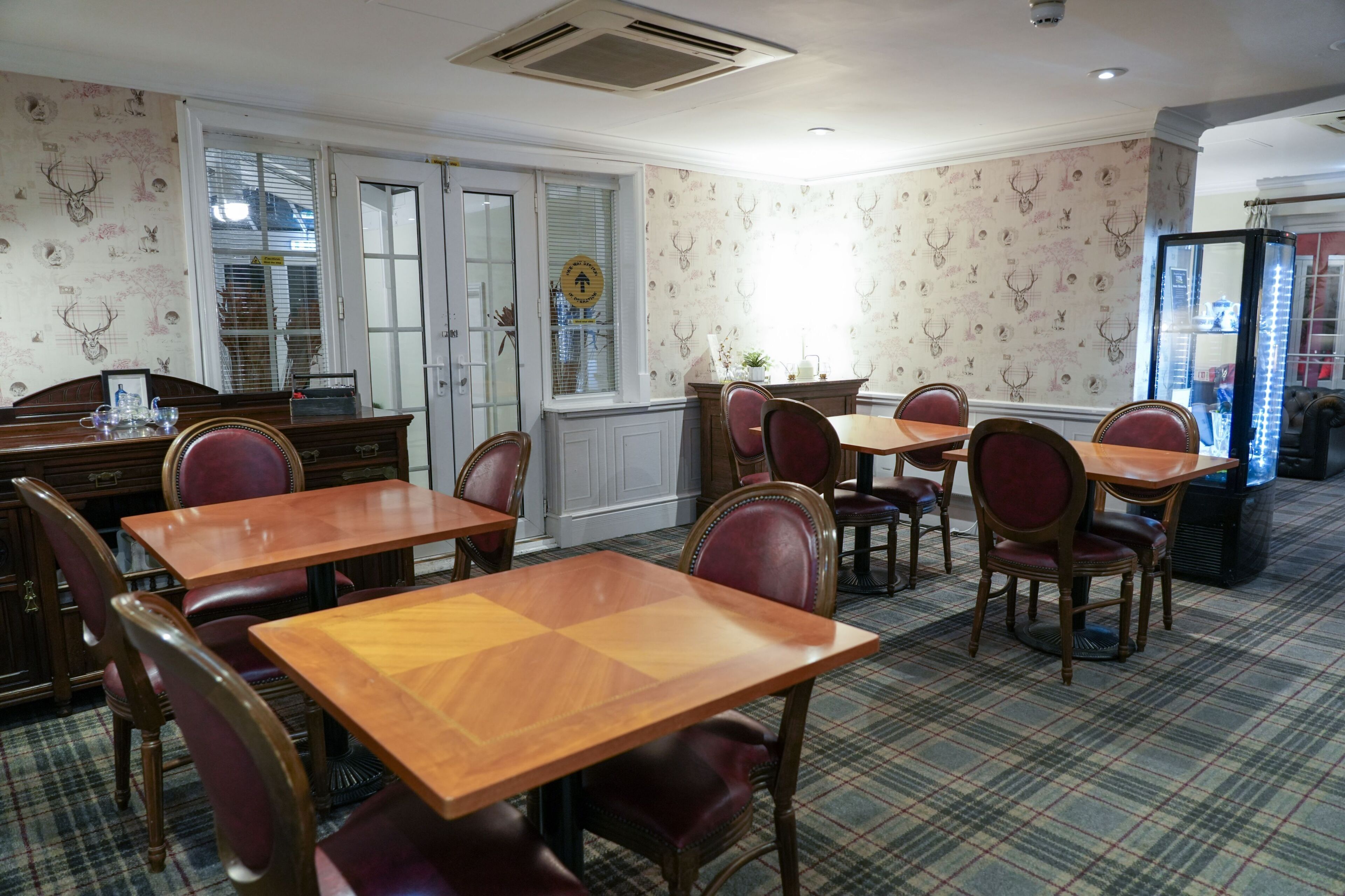 A dining area with wooden tables and red upholstered chairs, patterned wallpaper, and a plaid carpet.