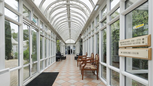 A covered corridor with glass walls, wooden chairs, and directional signs for rooms and the leisure club. - Basingstoke Hotel (Hook)