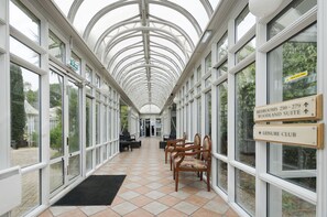 A covered corridor with glass walls, wooden chairs, and directional signs for rooms and the leisure club. - Basingstoke Hotel (Hook)