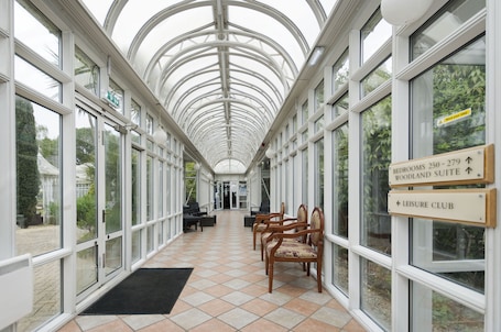 A covered corridor with glass walls, wooden chairs, and directional signs for rooms and the leisure club.