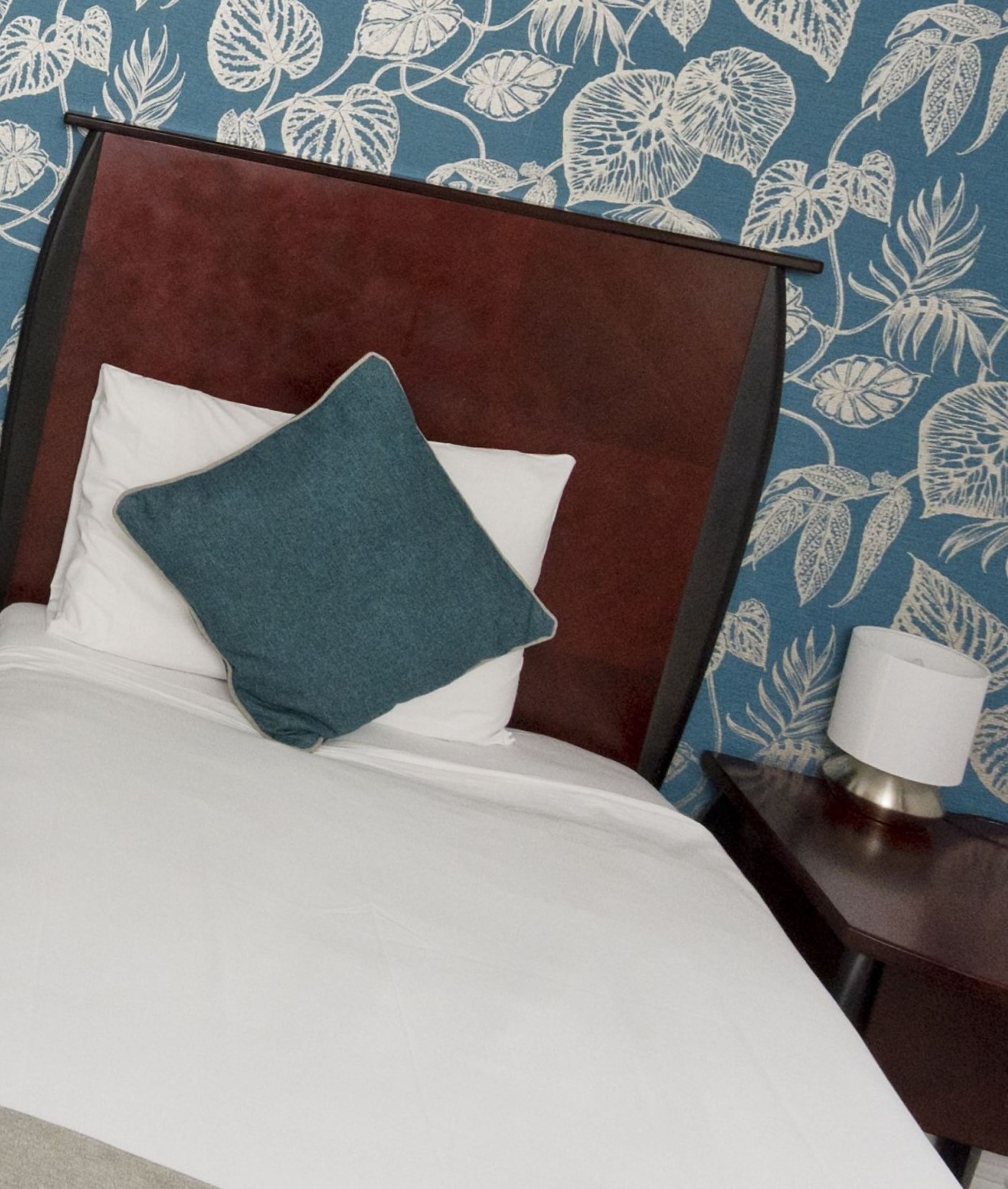 Una cama con cabecera oscura, almohadas blancas y una almohada color turquesa. Se ve una mesita de noche con una lámpara.