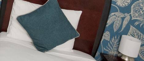 Una cama con cabecera oscura, almohadas blancas y una almohada color turquesa. Se ve una mesita de noche con una lámpara.