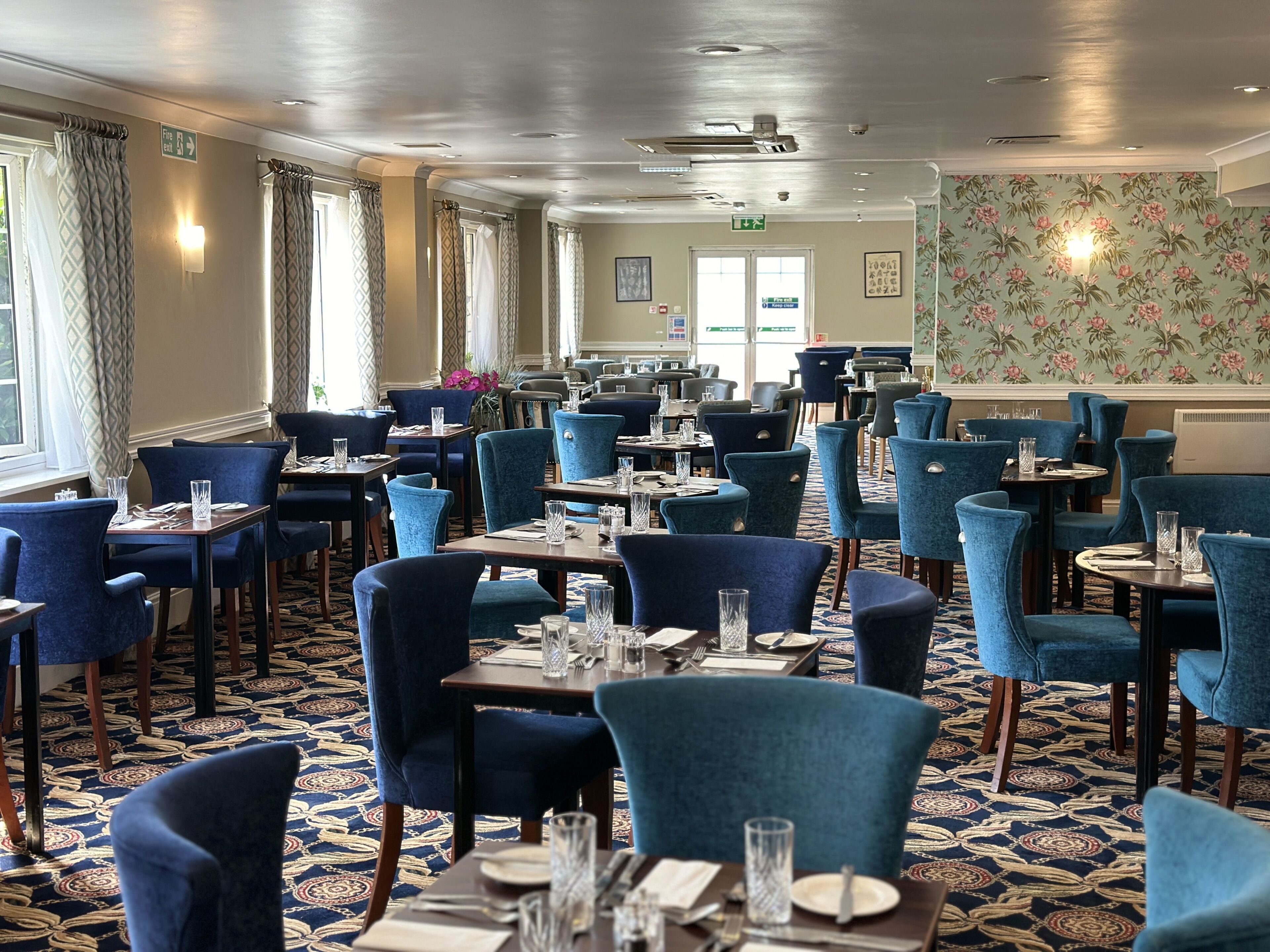 Une salle à manger d’hôtel avec des chaises à revêtement bleu, des tables dressées avec des couverts et des verres, une moquette à motifs et un papier peint floral.