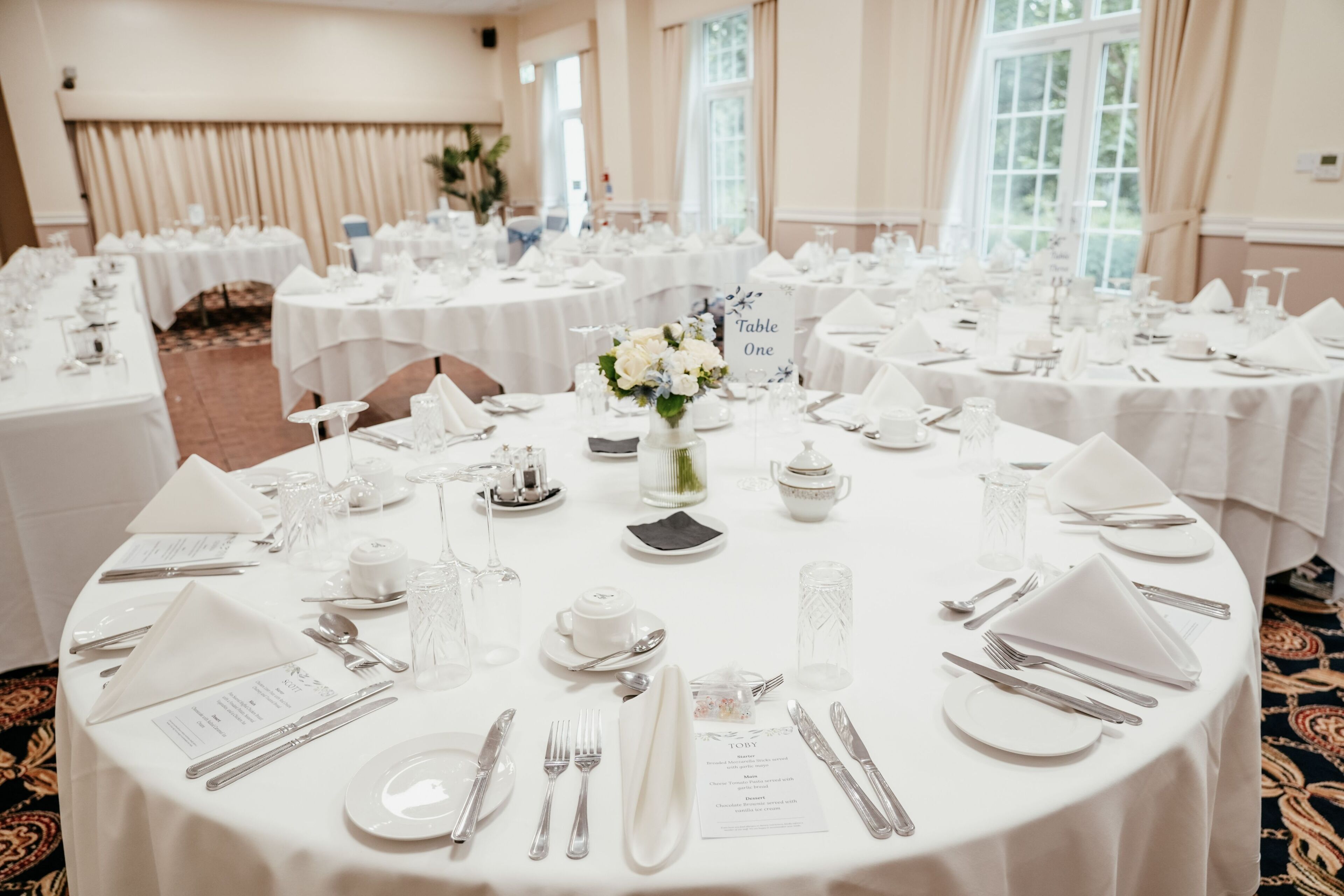 A banquet hall with round tables set for a formal event, featuring white tablecloths, silverware, and floral centerpieces.