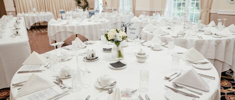 Un salón de banquetes con mesas redondas dispuestas para un evento formal, con manteles blancos, cubiertos de plata y centros de mesa florales.