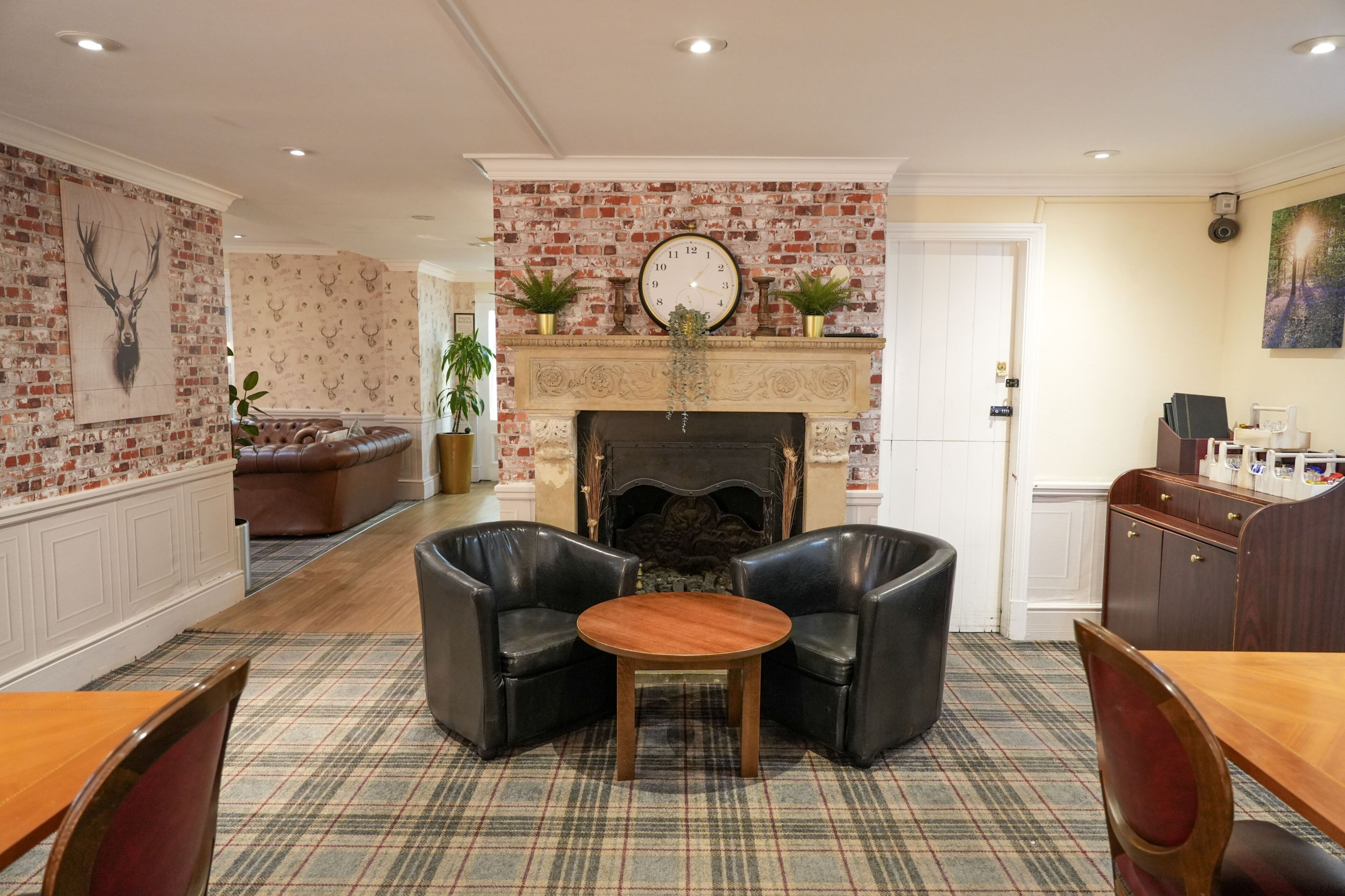 A cozy living room with a fireplace, leather armchairs, a plaid carpet, and a dining area with a wooden table and chairs.
