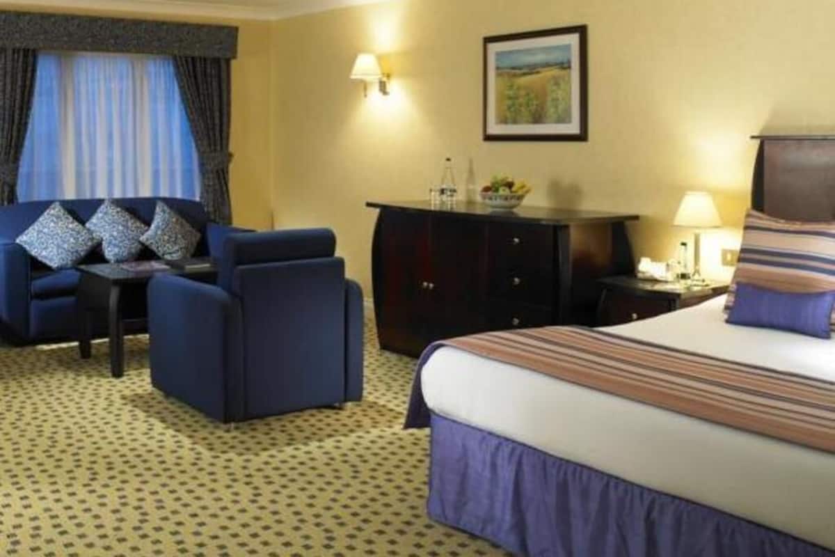 Une chambre d’hôtel comprenant un lit, deux fauteuils, un bureau, une commode et un tableau au mur.