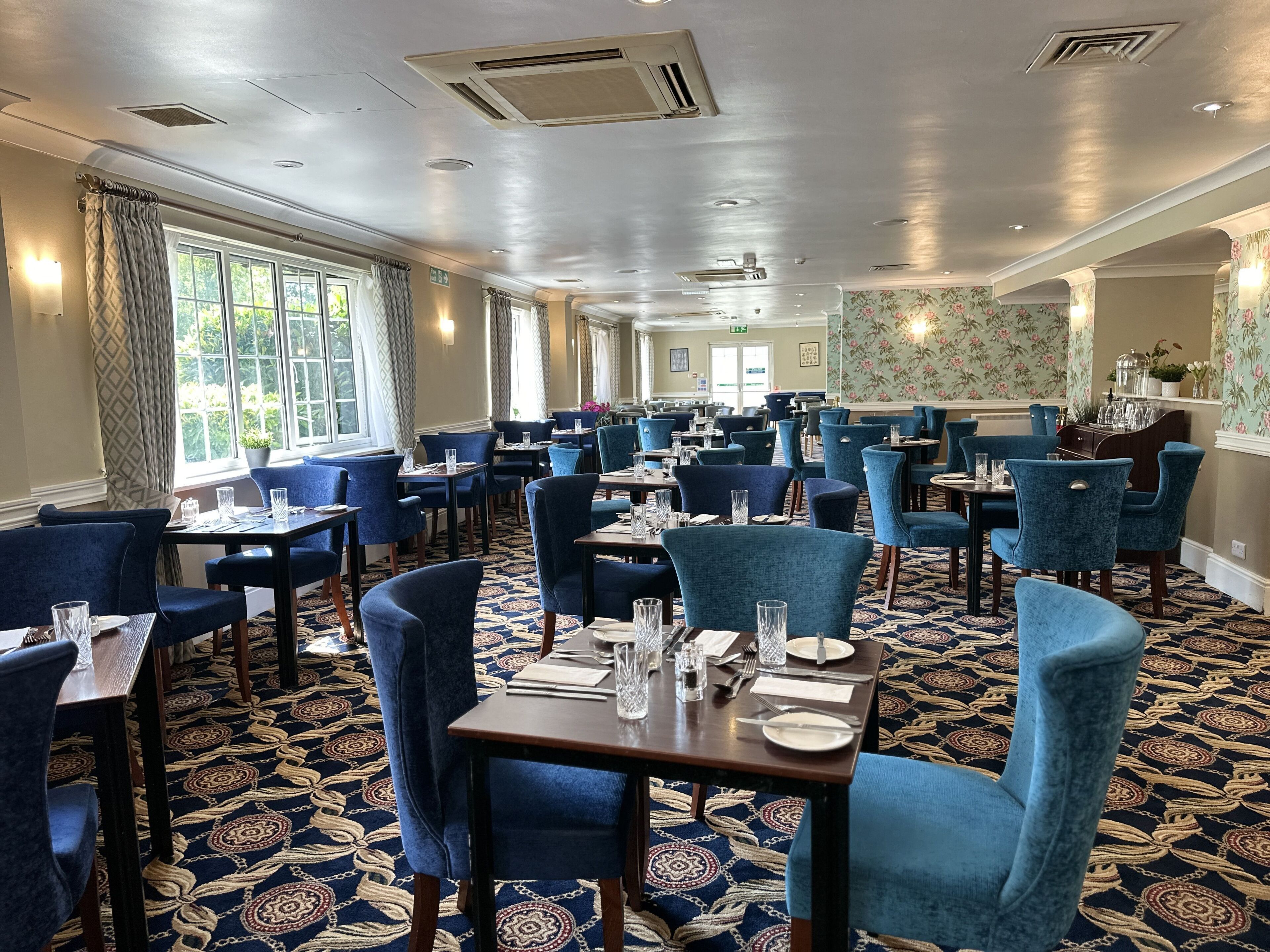 Een ruime restaurantruimte met hoge plafonds, grote ramen en blauwe gestoffeerde stoelen. De tafels zijn gedekt met borden, glazen en bestek.