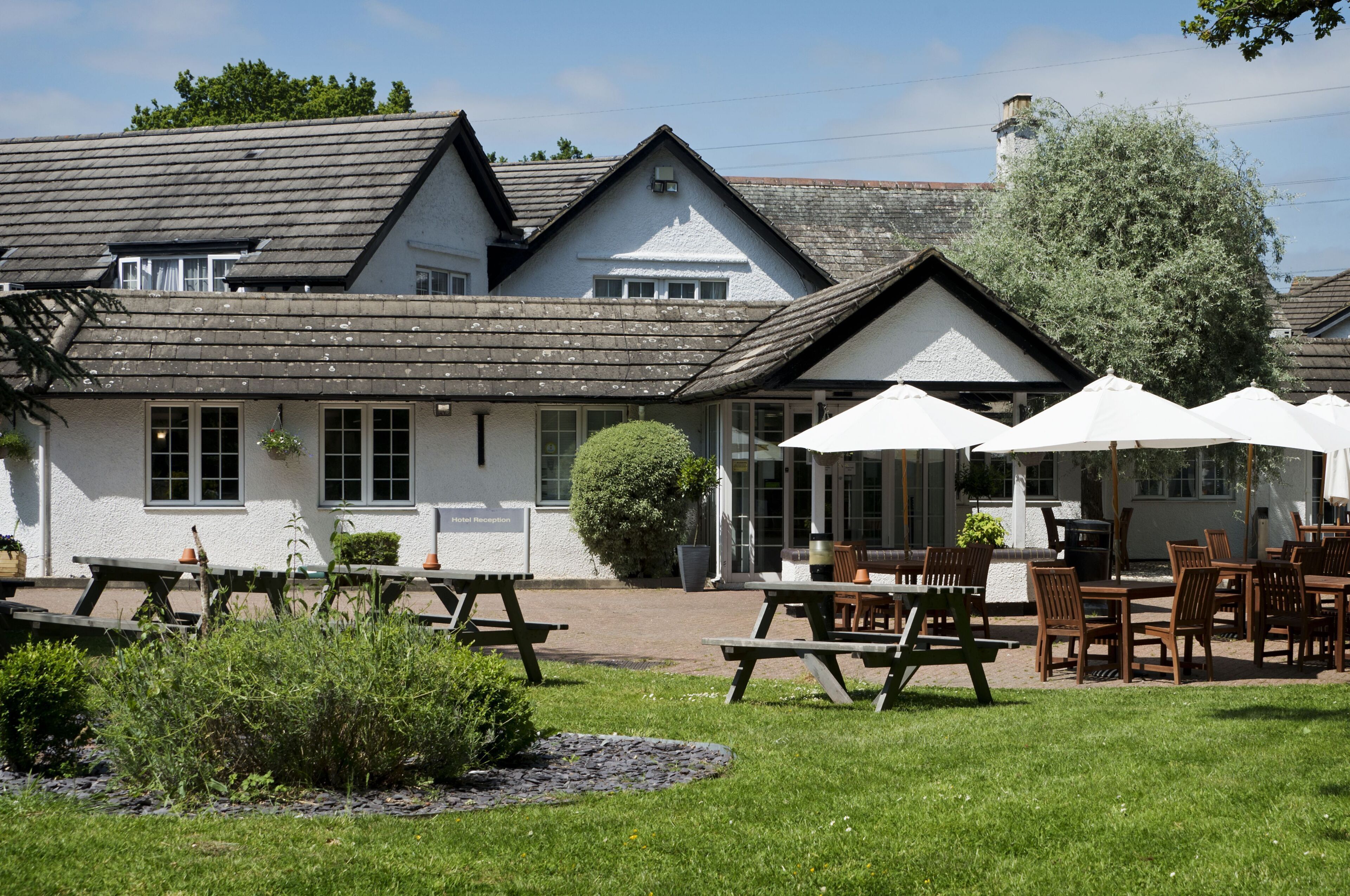A white building with a sloped roof, outdoor seating with umbrellas, and a garden area.