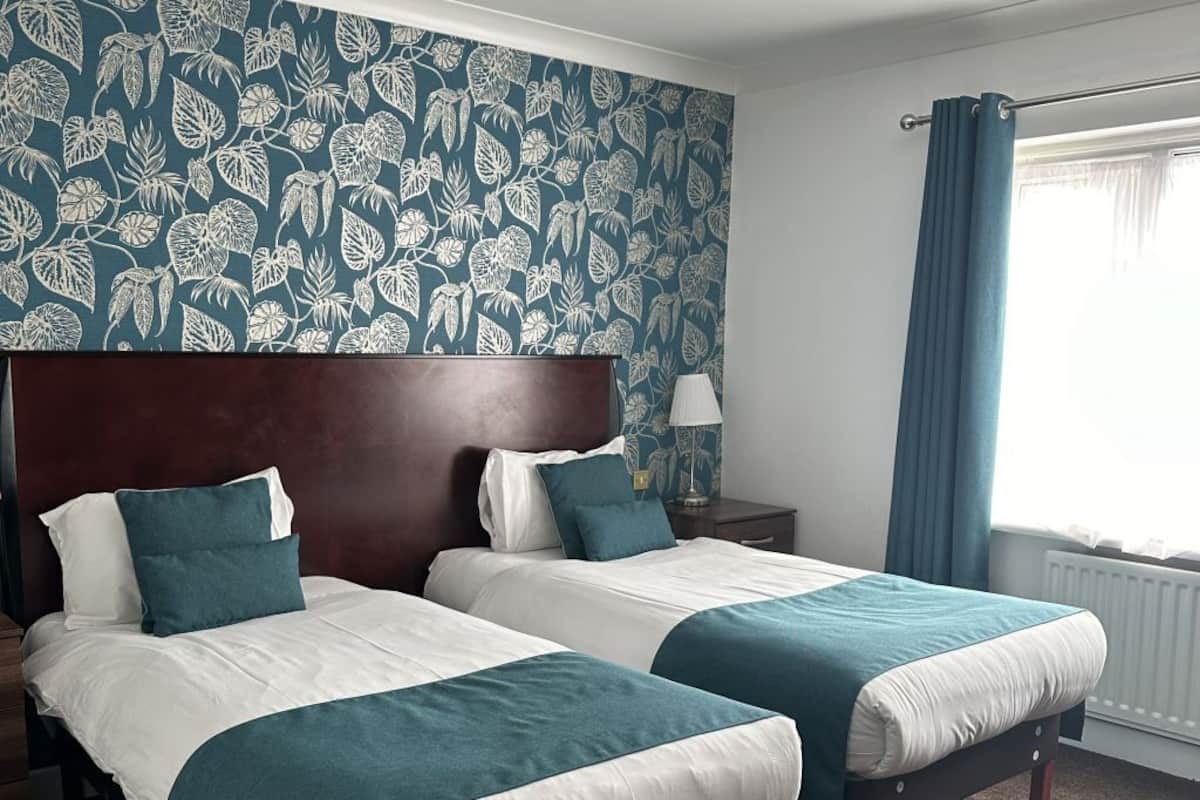Une chambre d’hôtel avec deux lits, une tête de lit en bois foncé, un papier peint à motifs, une table de chevet, une lampe et une fenêtre avec des rideaux.