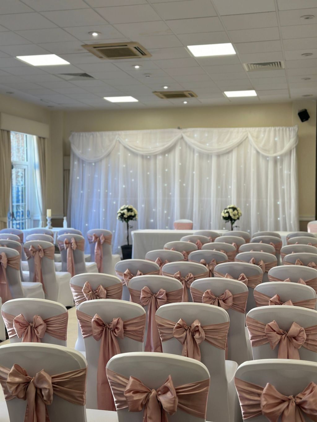 Rows of chairs with pink sashes and bows, a white draped backdrop, and a ceiling with recessed lighting.