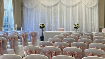 Rows of chairs with pink sashes and bows, a white draped backdrop, and a ceiling with recessed lighting.