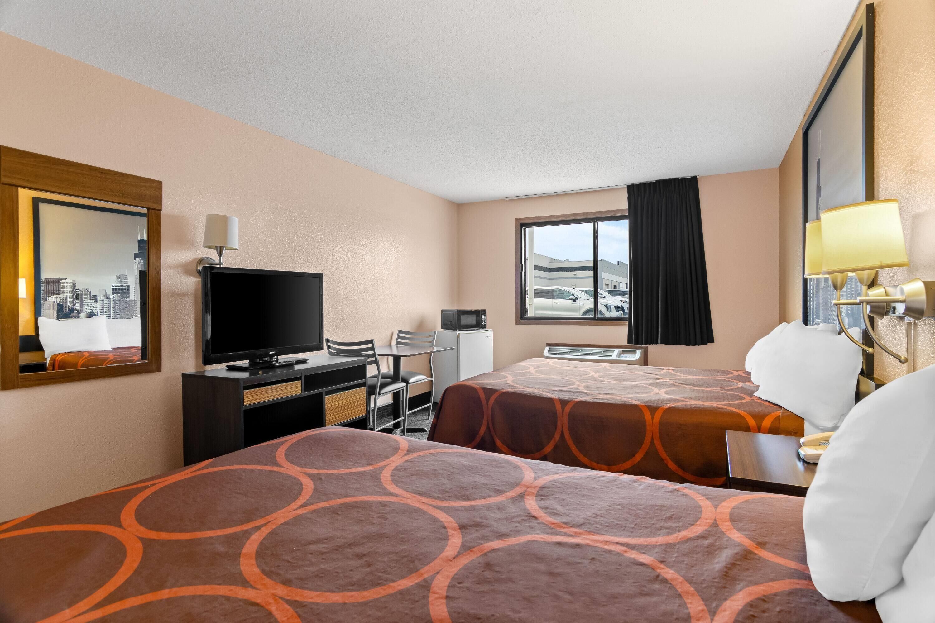 Habitación de hotel con dos camas, un escritorio con televisor, una silla, microondas y ventana con vistas a edificios.