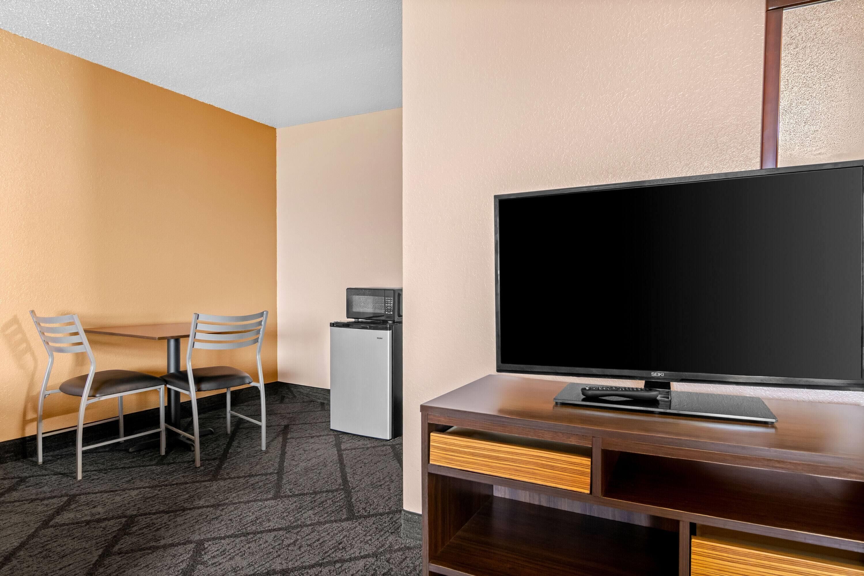 A hotel room with a TV, a small fridge, a desk with a chair, and a carpeted floor.