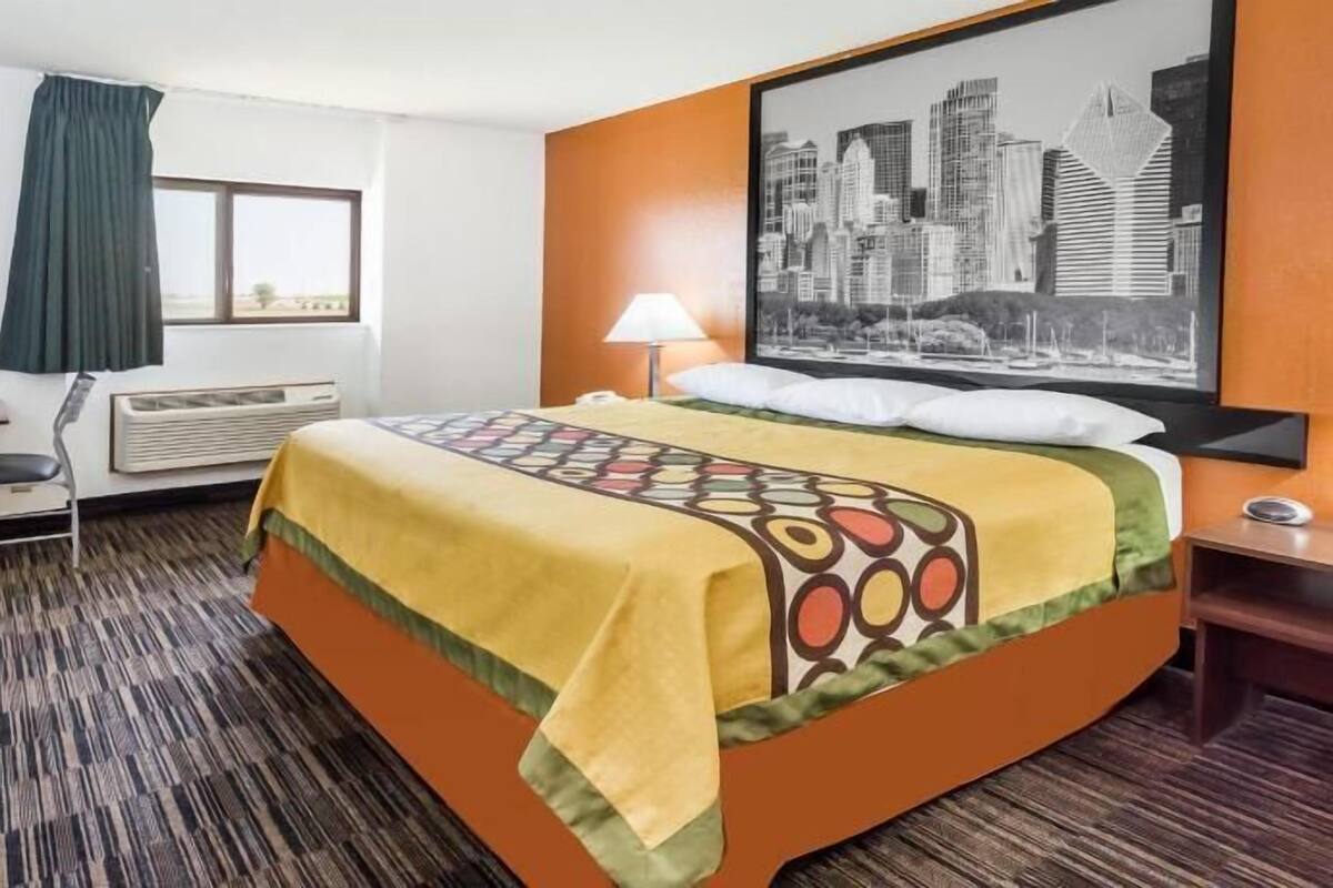 Une chambre d’hôtel équipée d’un lit, d’un bureau, d’une chaise, d’un climatiseur, d’une lampe et d’un tableau représentant un paysage urbain accroché au mur.