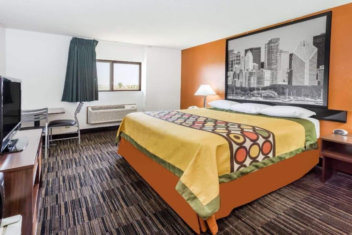 Une chambre d’hôtel comprenant un lit, un bureau, une chaise, un téléviseur, un appareil de climatisation, une fenêtre avec des rideaux et un tableau représentant un paysage urbain accroché au mur.