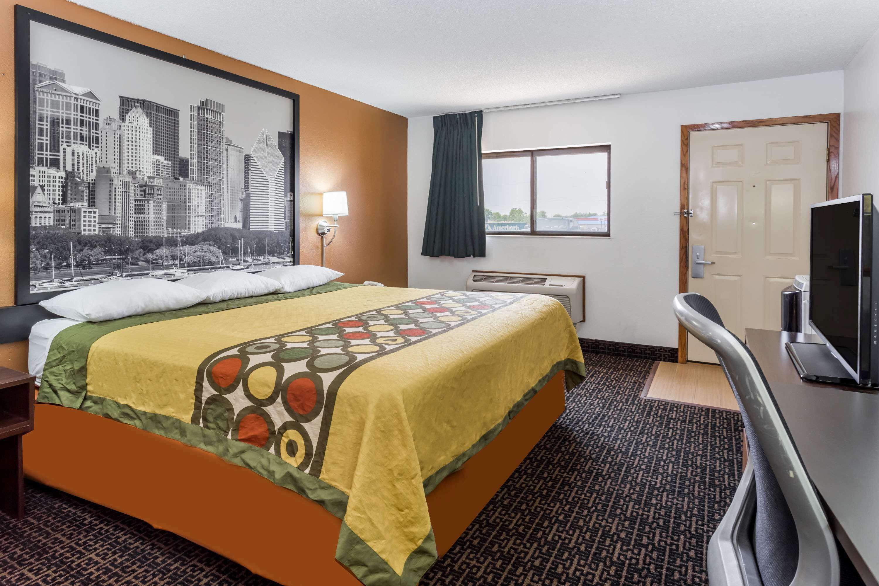Habitación de hotel con una cama grande, un escritorio con televisor, una ventana con vista a la ciudad y un cuadro de un paisaje urbano en la pared.