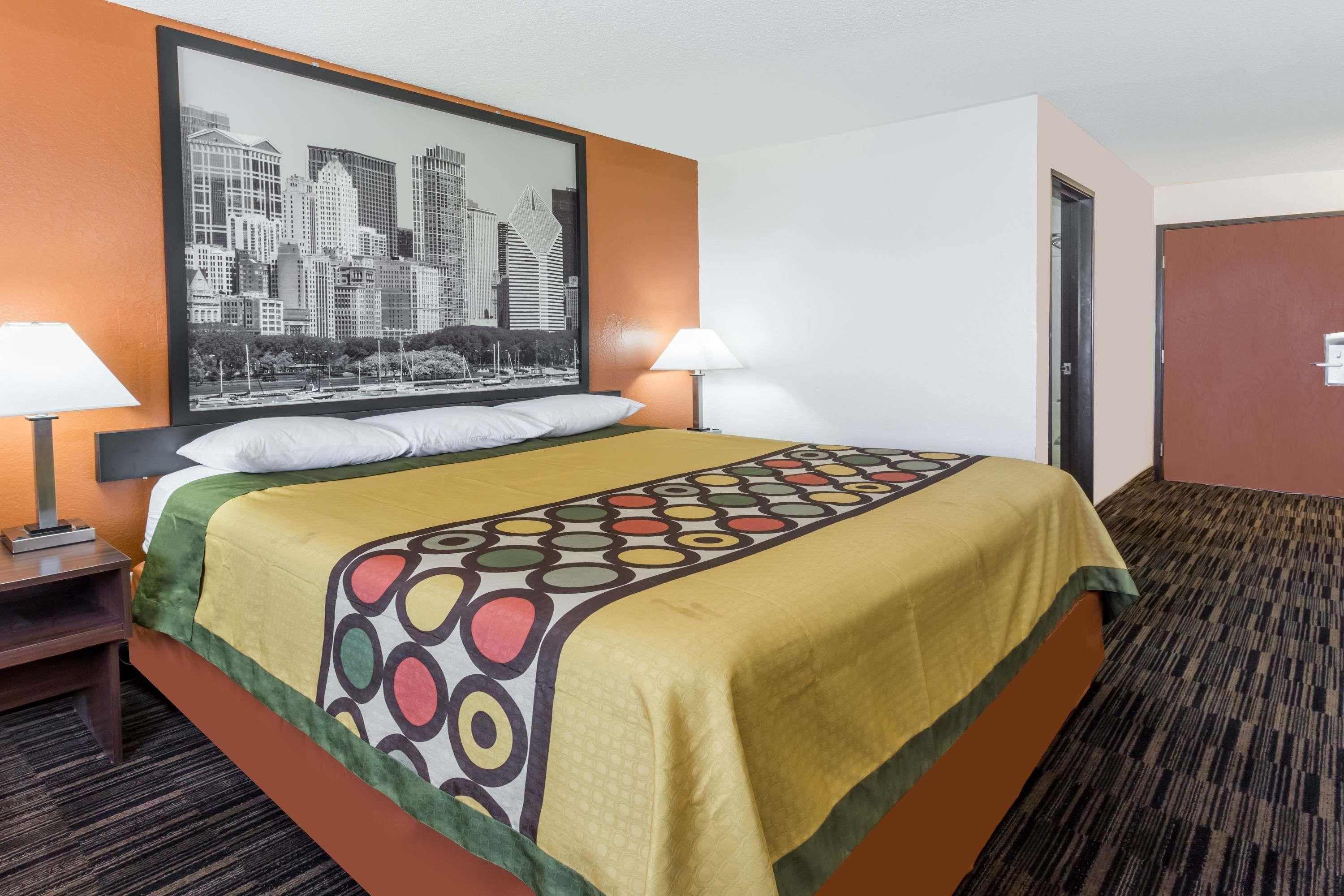 A hotel room with a bed, two bedside lamps, a cityscape artwork on the wall, and a door to the right.