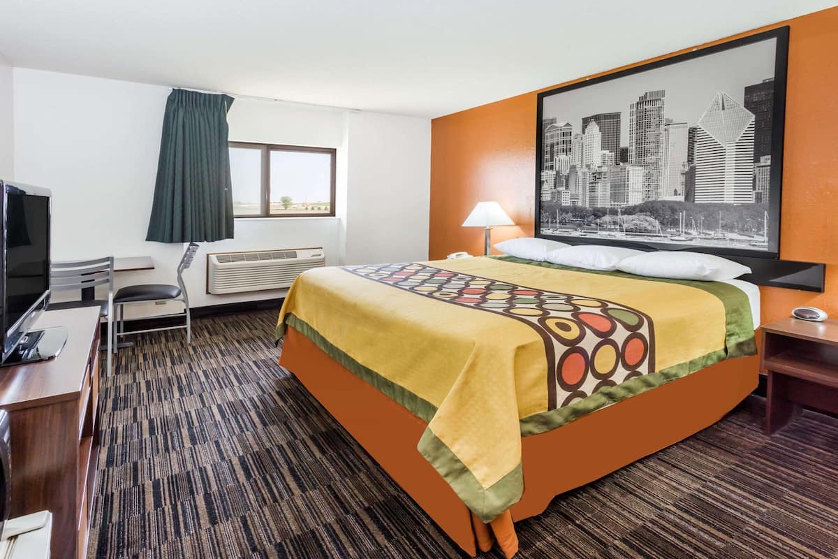 Une chambre d’hôtel avec un lit, un bureau avec un ordinateur, une chaise, une fenêtre avec des rideaux, un climatiseur, une lampe et un tableau représentant un paysage urbain accroché au mur.