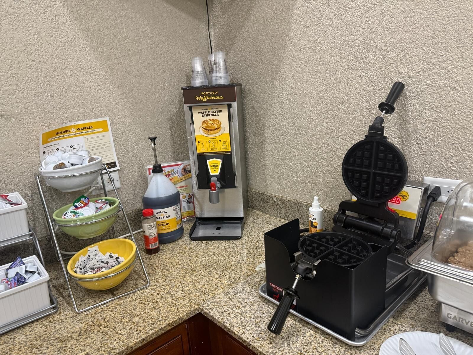 Un área de cocina con una plancha para waffles, dispensador de masa para waffles y diversos utensilios de cocina.