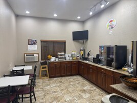 Una pequeña kitchenette con cafetera, refrigerador y microondas. Hay una ventana con persianas, un lavamanos y una pequeña mesa con sillas.