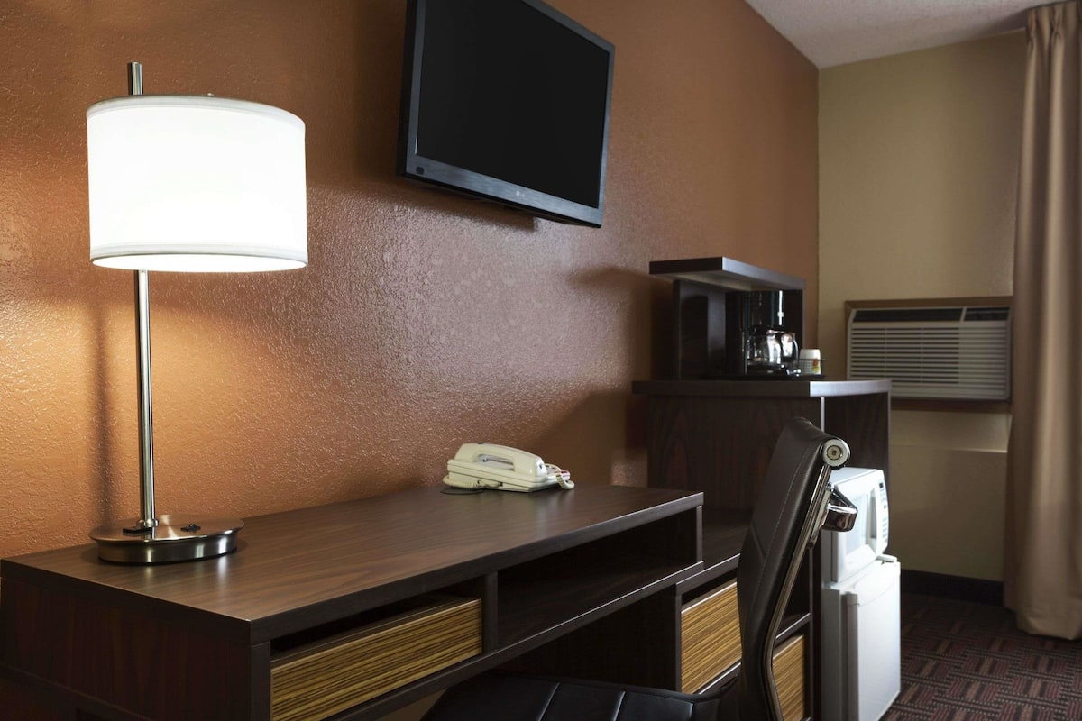 A hotel room with a desk, a lamp, a flat-screen TV, a telephone, a microwave, and a trash bin.