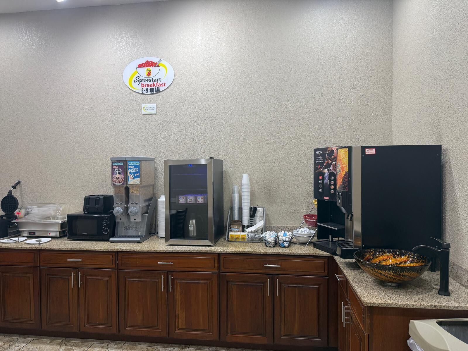 A breakfast area with a Superstart breakfast sign, a coffee machine, a refrigerator, and a bowl of fruit on the counter.