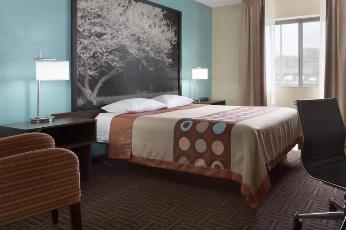 A hotel room with a bed, a desk, a chair, a large painting on the wall, and a window with curtains.