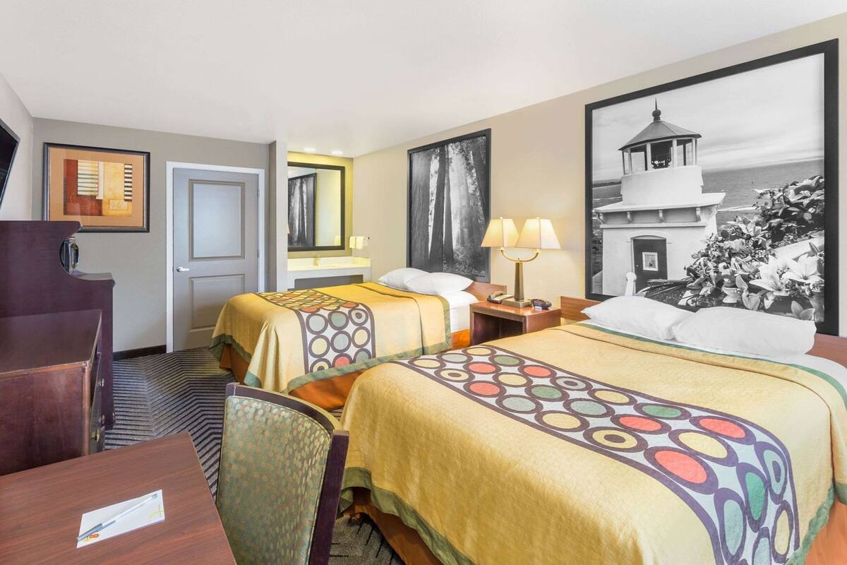 A hotel room with two beds, a desk, a chair, a lamp, and a large framed picture on the wall.