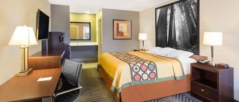 Quarto de hotel com cama, escrivaninha, cadeira, televisão, espelho, pintura e mesa de cabeceira com abajur.