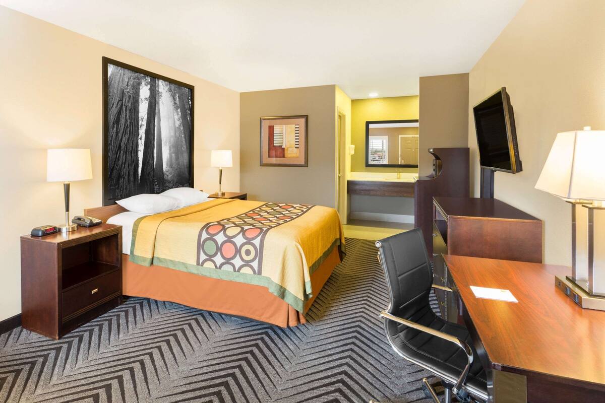 A hotel room with a bed, a desk with a chair, a television, and a bathroom visible in the background.